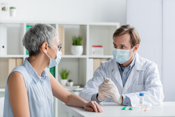 Doctor in medical mask sitting near mature patient and vaccine with pills during consultation