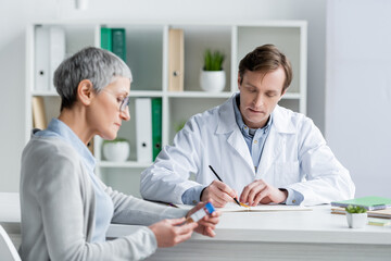 Doctor writing on notebook near patient holding vaccine on blurred foreground