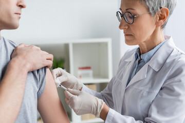 Mature doctor in eyeglasses doing vaccination of man on blurred foreground in hospital