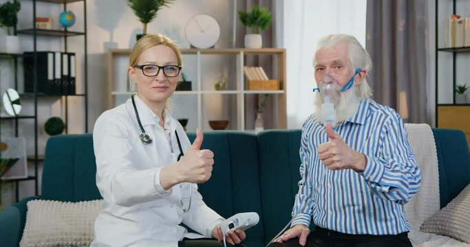 Senior grey-haired bearded man in inhaler and attractive smiling professional skilled female doctor looking at camera with thumbs up