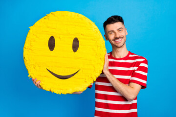 Photo of cheerful young happy man hold big face pinata smile emoji isolated on blue color background