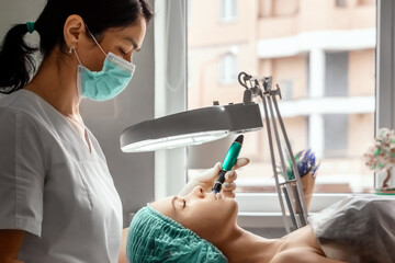 A beautician wearing medical gloves cleans the pores on the face using special equipment while...