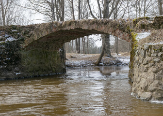 Fototapeta na wymiar a continuous boulder stone bridge with a brick used for masonry, early spring, bare trees, snow plan on the ground, Stone arch bridge over the river Kuja, Madona, Latvia