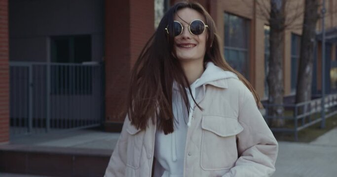 Cheerful young lady with brown hair taking fun while walking on city street. Pretty woman in sunglasses and casual wear spending free time with enjoyment. 