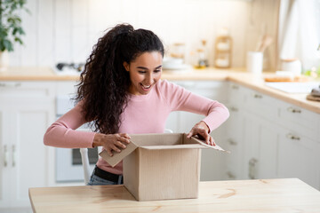 Excited happy young woman unboxing cardboard parcel In kitchen
