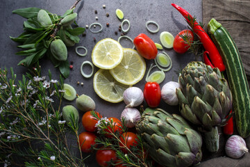 Colorful photo of fresh vegetables on a table. Cherry tomatoes, artichokes, lemon slices, leeks, pepper, zucchini and green almonds top view photo. Gray textured background. Healthy eating concept. 
