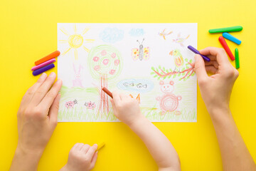 Mother and baby hands holding wax crayons and drawing colorful draw on paper on yellow table...