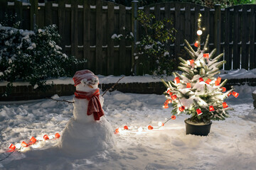 A potted Christmas tree and snowman stand in a snowy backyard at night.