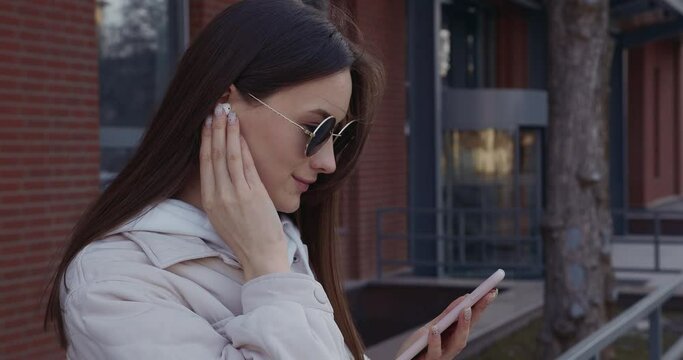 Adorable woman with smartphone in hands listening music in wireless earphones. Young brunette in sunglasses standing on street and using modern gadgets. 