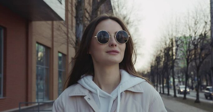 Adorable young brunette in sunglasses and casual outfit using modern smartphone while walking on city street. Concept of people, technology and lifestyle. 