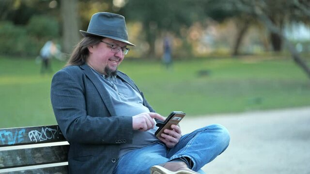 [4k] long haired goatee best ager sitting in park playing on his smartphone wearing hat jacket and glasses