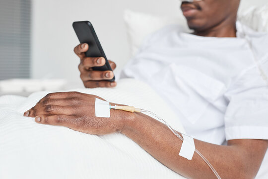 Close up of African-American man in hospital bed and using smartphone with focus on iv drip catheter in hand, copy space