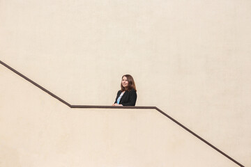 A woman with beautiful brown hair in a black jacket stands at the railing on the stairs leading up. The wall of a large bright yellow house with dark brown stair railings.