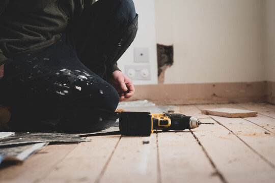 A workman drills a hole on the floor of a house indoors in preparation for laying out new floorboards. 