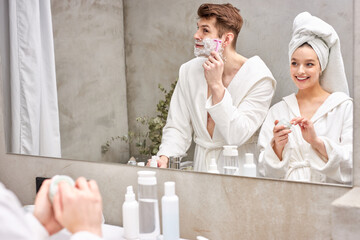 attractive female in bathrobe and towel use moisturizing cream while boyfriend shaving stubble