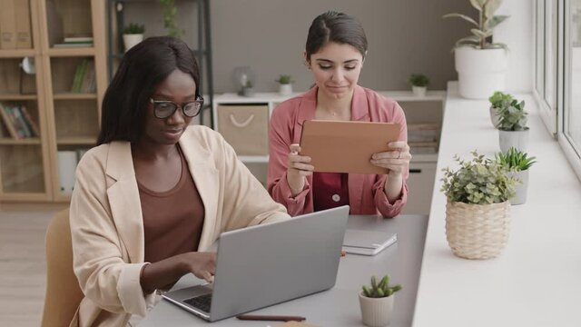 Lockdown of young African businesswoman sitting at desk in office, working on laptop while her female mixed-race colleague sitting nearby and using digital tablet