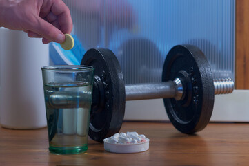 Classic dumbbell with a glass of water and a set of pills. Male hand puts a effervescent tablet vitamin pill into a glass.
