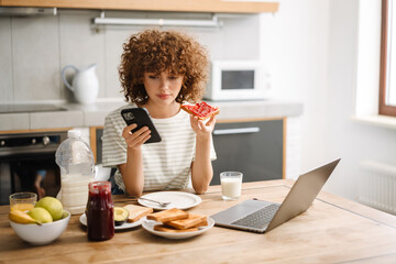 Curly young woman using mobile phone and laptop while having breakfast - obrazy, fototapety, plakaty