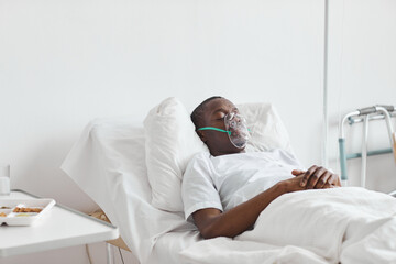 Portrait of African-American man sleeping on hospital bed with oxygen mask in white room, copy space