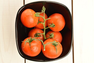 Several ripe, bright red, organic, delicious cocktail tomatoes in a black ceramic plate, close-up, on a table made of white painted natural wood.