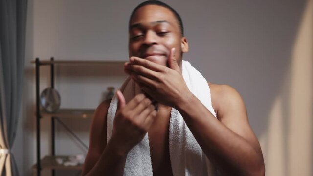 Male skincare. Grooming routine. Positive lifestyle. Relaxed happy shirtless athletic African man with towel enjoying dancing singing touching face after shower smiling.