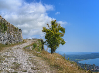 Rocky path in the mountains along the walls of the old fortress on a summer day. An old lonely tree is soloing against a blue sky with textured white clouds. Below the mountain is a blue sea 
