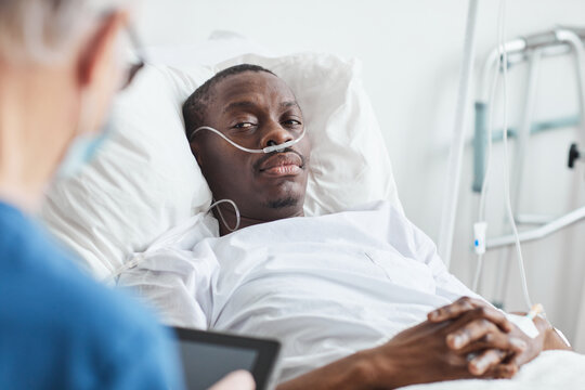 Portrait of African-American man lying in hospital bed and looking at doctor, copy space