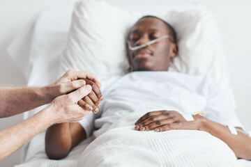 Close up of caring relative holding hand of African-American man lying in hospital bed, copy space