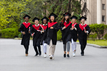 Fototapeta na wymiar Cheerful multiracial students in graduation costumes walking by university campus