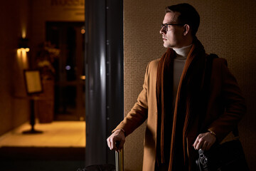 Side view portrait of male in eyeglasses standing with luggage, waiting for someone in hotel hall