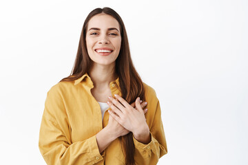 Beautiful and tender woman smiling, holding hands on heart, care for you, being touched deeply, looking heartfelt and thankful, thank you, standing over white background