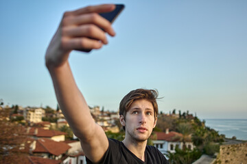 Fototapeta na wymiar Young white man takes photo of himself against backdrop of an old town in Antalya using smartphone.