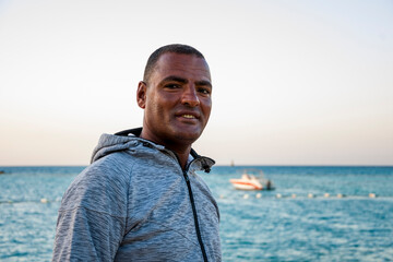 Portrait of a dark-skinned Egyptian man of 40 years in a hoodie against the background of the sea and a boat.