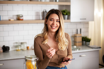 Young woman in kitchen. Beautiful woman using phone while making food.