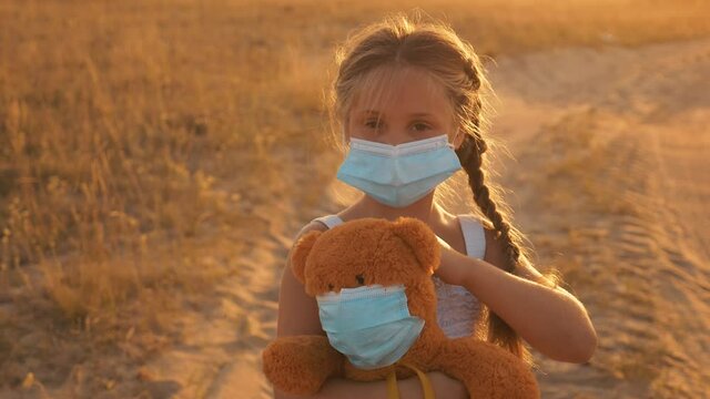A child stands on the field with a soft toy in his hands. A child in a medical mask isolated during the quarantine of the virus. A kid plays doctor with a Teddy bear. The concept of health care.