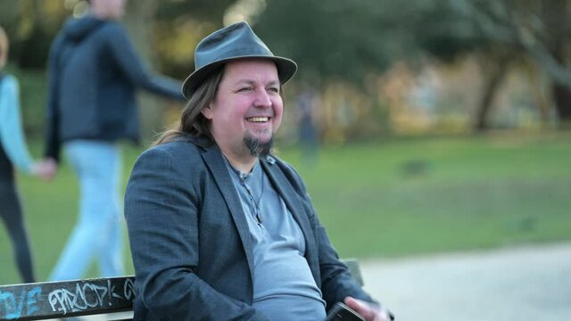 [4k] long haired old rocker is happy sitting on park bench putting on head
