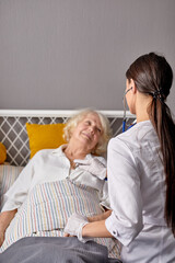 Obraz premium rear view on helpful female nurse checking sounds in heart and lungs of sick senior woman lying on bed
