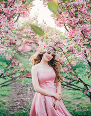 Spring happy woman walks in blooming sakura flowers garden. Medieval girl, blonde lady. pink silk dress. fairy image art photo. Blond long hair fly fluttering in wind, wreath crown. Smiling cute face.