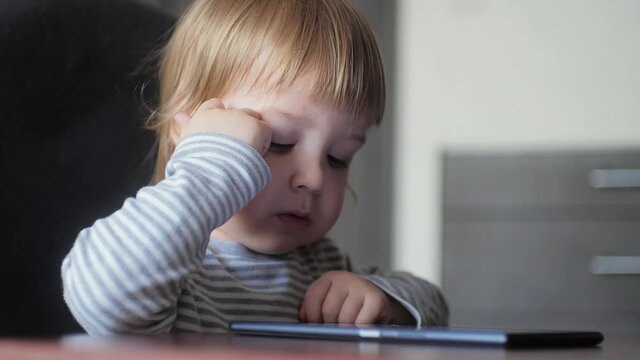 Portrait of a child sitting at a desk and enthusiastically looking and tapping the screen of a digital tablet. Modern electronic devices for the development of children.