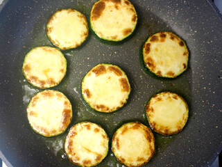 the process of frying green sliced zucchini slices in vegetable oil in a gray frying pan top view . vegetarianism
