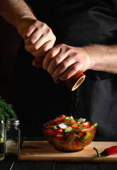 Chef in restaurant kitchen adds peppers to a fresh vegetable salad. Close-up of a cook hands holding mill. Cooking healthy and tasty food