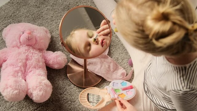 Little child girl paints eyes with eyeshadow brush, plays makes makeup in front of the mirror in the children's room. Imitates adults making experiments with cosmetics. Role-playing games.