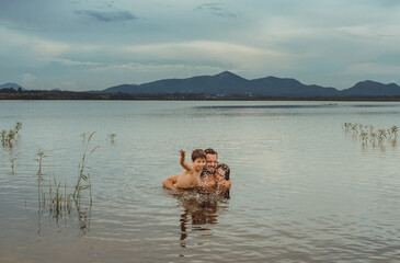 Obraz na płótnie Canvas A man hugging two kids who splash water into a calm lake, at Iraí Reservoir, in the city of Pinhais, State of Paraná, Brazil. In the background, the mountains that form the 
