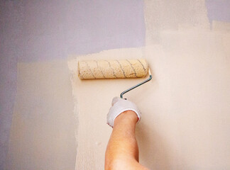 mans hand in cloth glove with paint roller applies fresh paint and primer to wall surface. process of applying water-based paint and primer with paint roller on surface for  interior design of room.