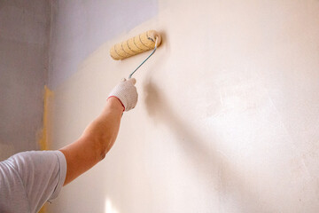 mans hand in cloth glove with paint roller applies fresh paint and primer to wall surface. process of applying water-based paint and primer with paint roller on surface for  interior design of room.