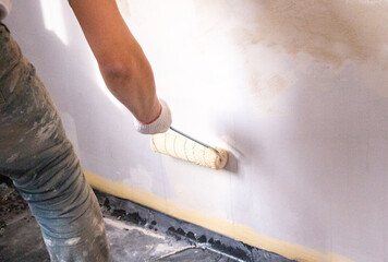 mans hand in cloth glove with paint roller applies fresh paint and primer to wall surface. process of applying water-based paint and primer with paint roller on surface for  interior design of room.