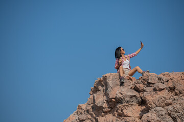 woman hiking in mountains standing on rocky summit ridge with backpack and pole looking out over landscape, happy female making self portrait in mountains, holding smartphone camera to take picture.