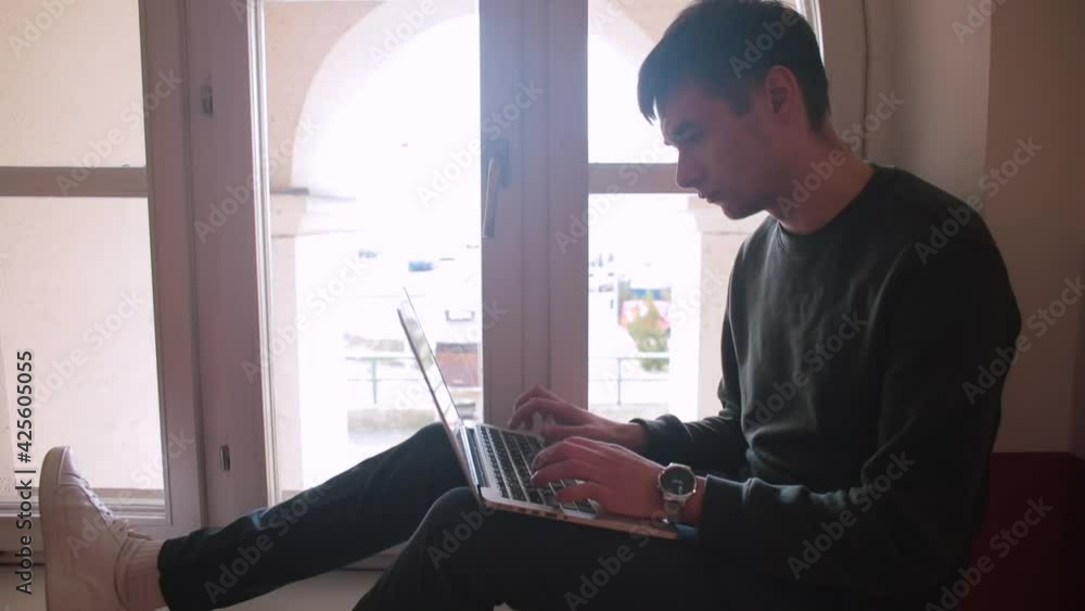 Poster focus on the screen. light in the window. a man uses a laptop while sitting at the window. he does r
