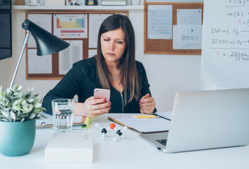 Serious female professor having break with online class and using her mobile phone to send text message. Teacher is working at home and video conferencing using her laptop, sitting and holding phone