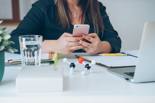 Cropped photo of unrecognizable female chemistry teacher sitting at desk and holding mobile phone. Young professor or student having break with online class and using smartphone to send text message